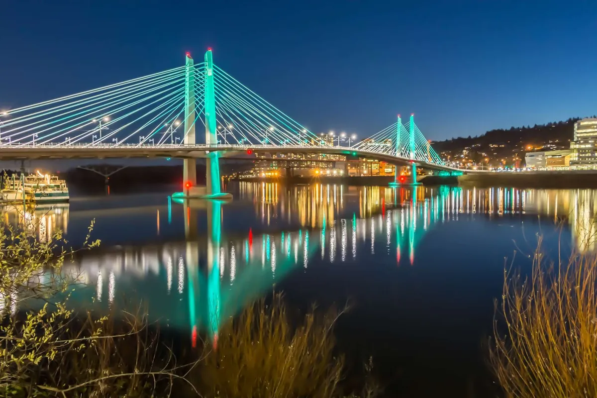 A scenic evening shot featuring a bridge with its reflection shimmering on the water, framed by city lights. The tranquil, romantic atmosphere evokes the allure of evening strolls, urban exploration, and picturesque downtown experiences.