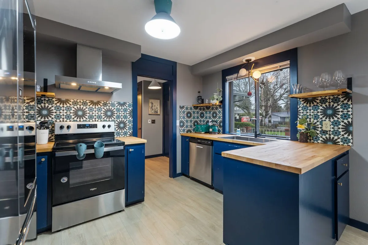 A vibrant kitchen featuring a colorful tile backsplash, wood countertops, blue vintage-style cabinetry, and a large window that fills the space with natural light. Modern appliances are visible alongside ample counter space, blending retro charm with convenience.