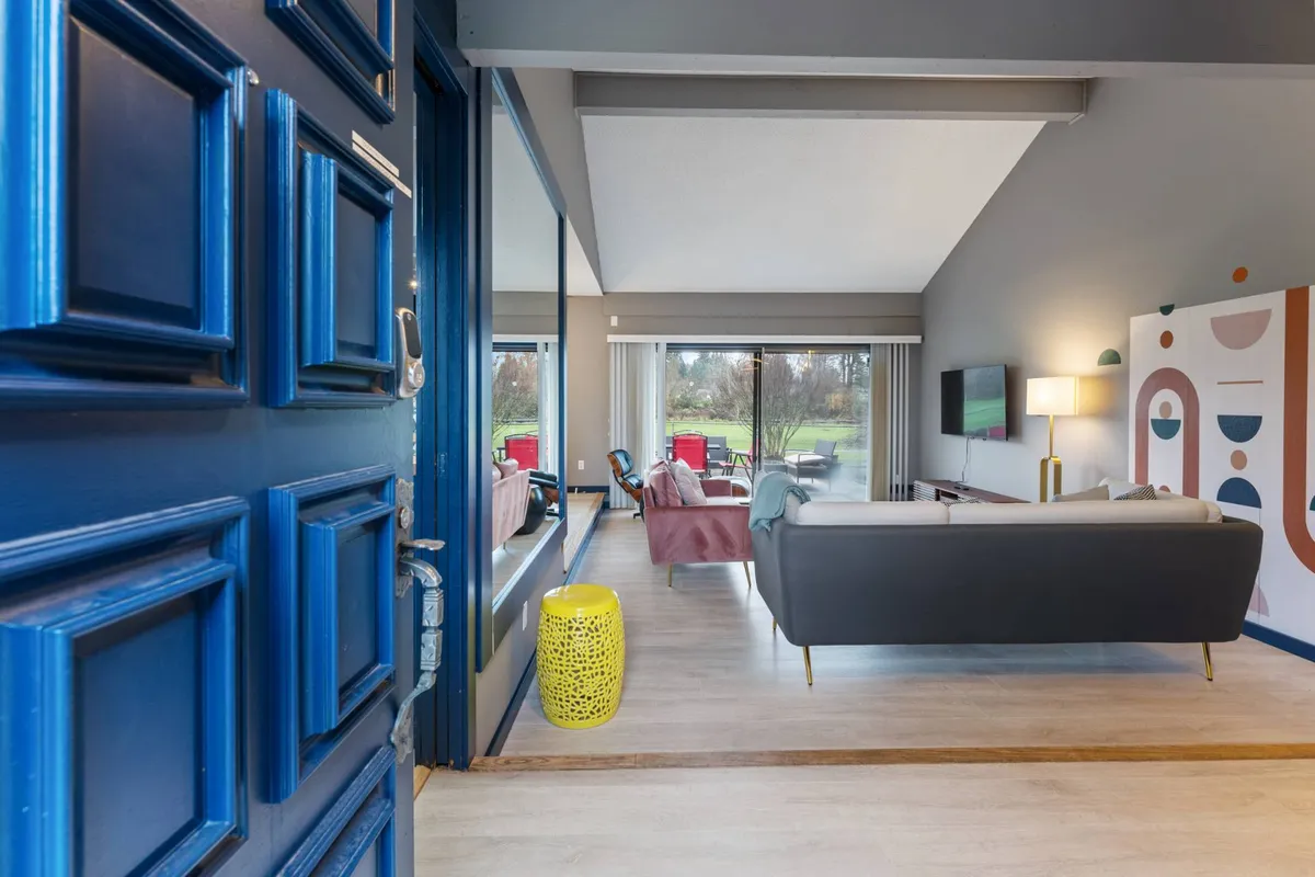 This cheerful, sunlit living area centers on a blue door, yellow stool, red chairs, and large windows that let in natural light. Artistic and welcoming, the space is perfect for casual gatherings, sunny brunches, or cozy evenings, capturing the retro spirit of the 1970s while maintaining a spacious, open feel.