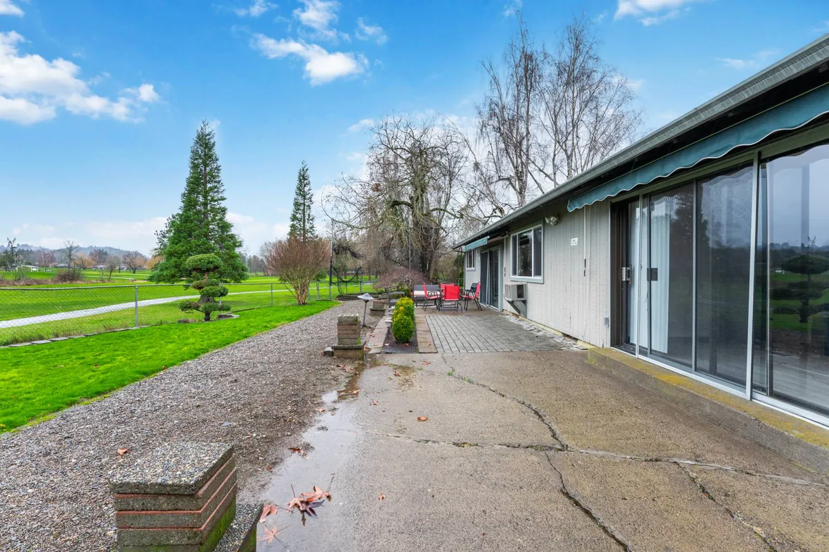 A peaceful backyard scene featuring a green lawn, cozy outdoor seating, a backdrop of mature trees, and a pathway leading through the landscape. The space feels connected to nature and is ideally suited for sipping coffee, watching golfers, or enjoying quiet moments in the sunshine.