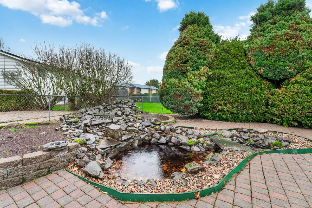 A tranquil exterior view of Golfview Ranch’s landscaped backyard, featuring a rock pond, lush bushes, and a winding stone pathway. The space is inviting, well-maintained, and perfect for outdoor relaxation or small gatherings. The serene setting showcases the property’s peaceful atmosphere.