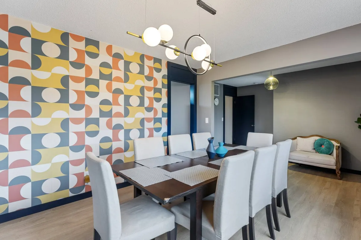 A cozy dining nook featuring a dining table set against geometric wallpaper, a stylish lighting fixture overhead, and an accent chair. The scene is set for intimate dinners, brunches, or lingering coffee conversations.