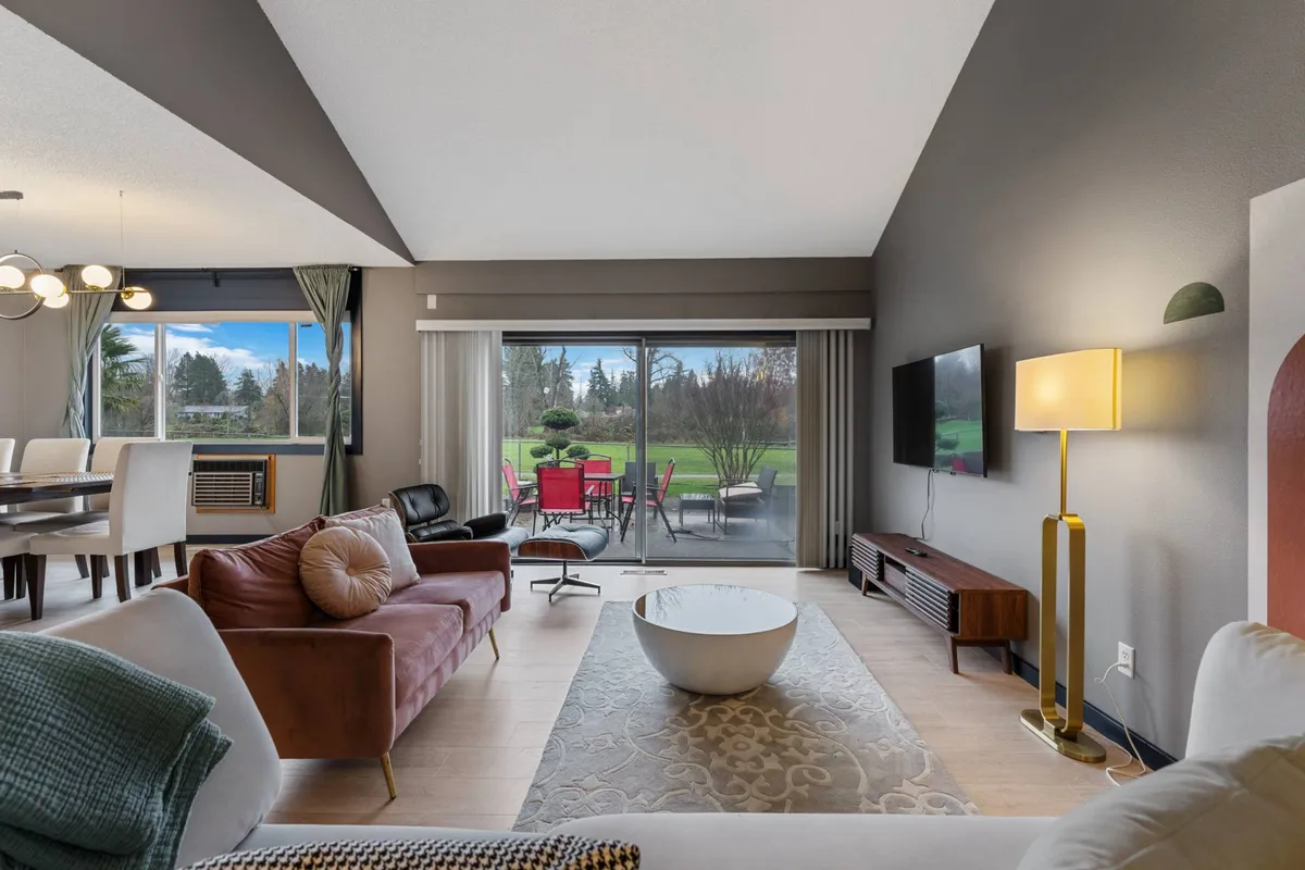 This image captures a welcoming, open-concept living space featuring a stylish sofa, modern round coffee table, and a dining area visible in the background. Large windows flood the room with natural light, offering a view of the outdoors and emphasizing the spacious, interconnected layout. The decor nods to mid-century and retro styles, complementing the 1970s charm mentioned in the section.