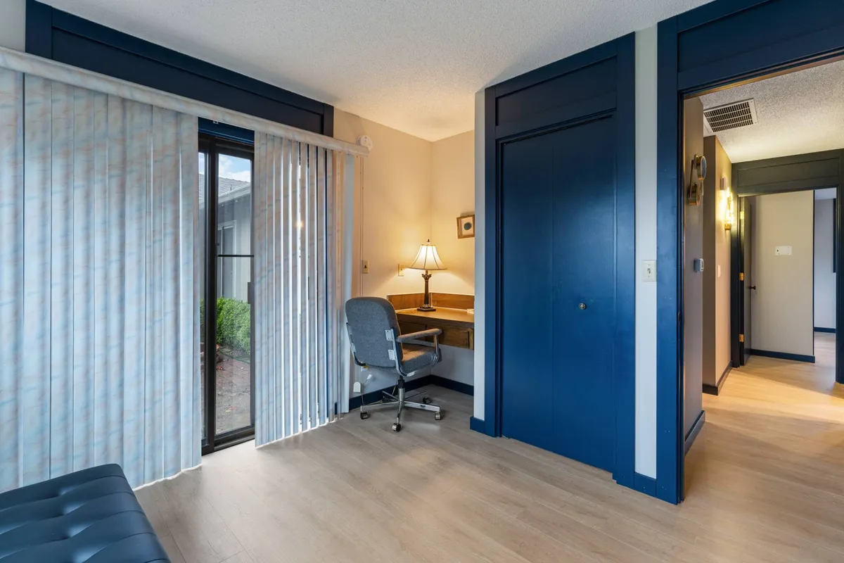 A dedicated workspace featuring a desk with a lamp, a sliding glass door, and a comfortable chair. The area is bright and positioned for productivity, making it suitable for remote work or study. Natural light streams in, suggesting a pleasant environment for extended work hours.