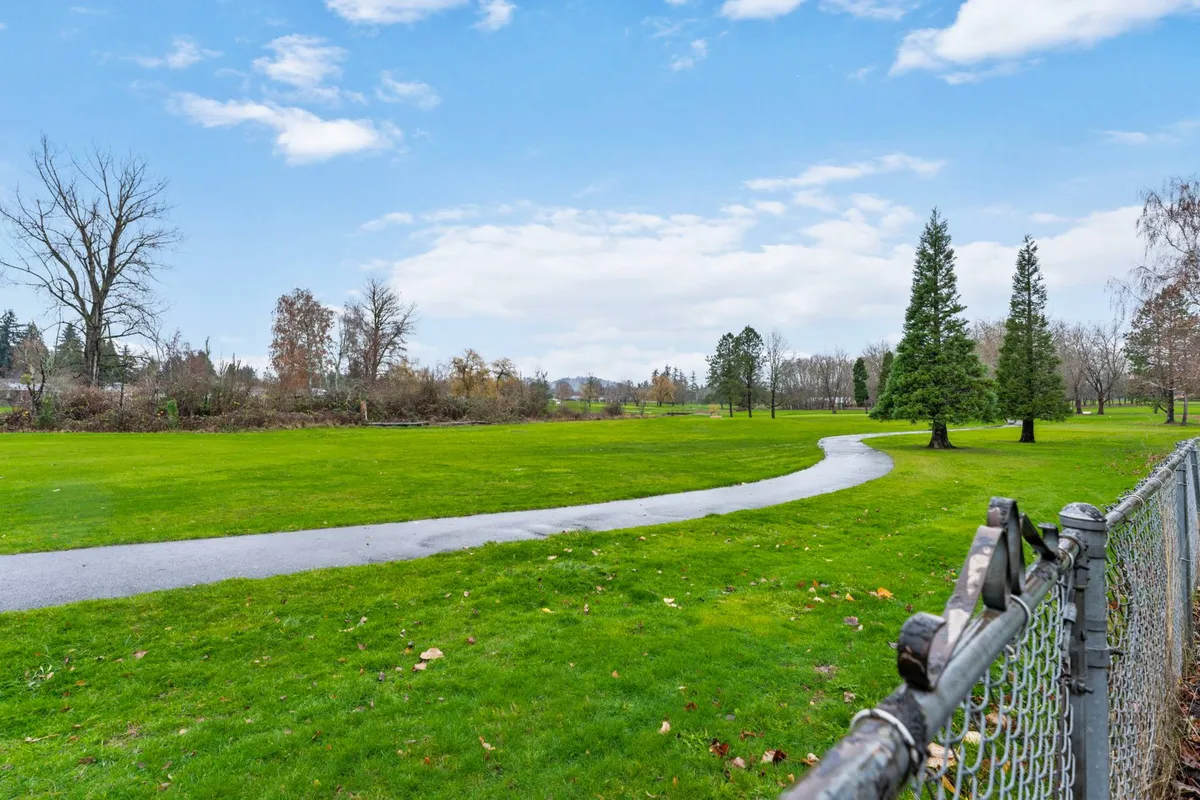 A lush, green park-like area with a winding path through tall evergreens and open grassy spaces, bordered by a sturdy fence. The scene invites outdoor exploration and activities like walking, jogging, and picnicking.