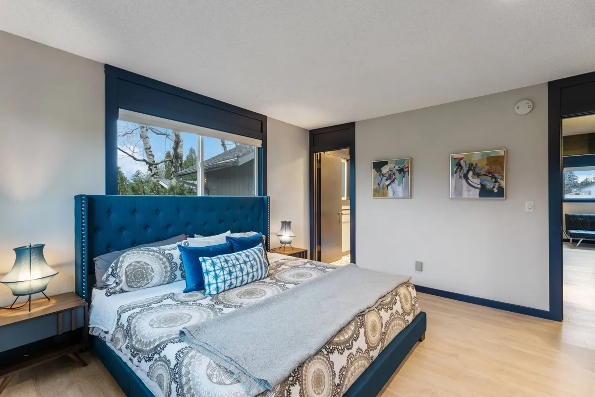 This image presents a serene bedroom with a blue headboard, decorative pillows, lamps on nightstands, artwork on the walls, and large windows that fill the space with natural light. The room feels inviting and restful, encouraging relaxation.