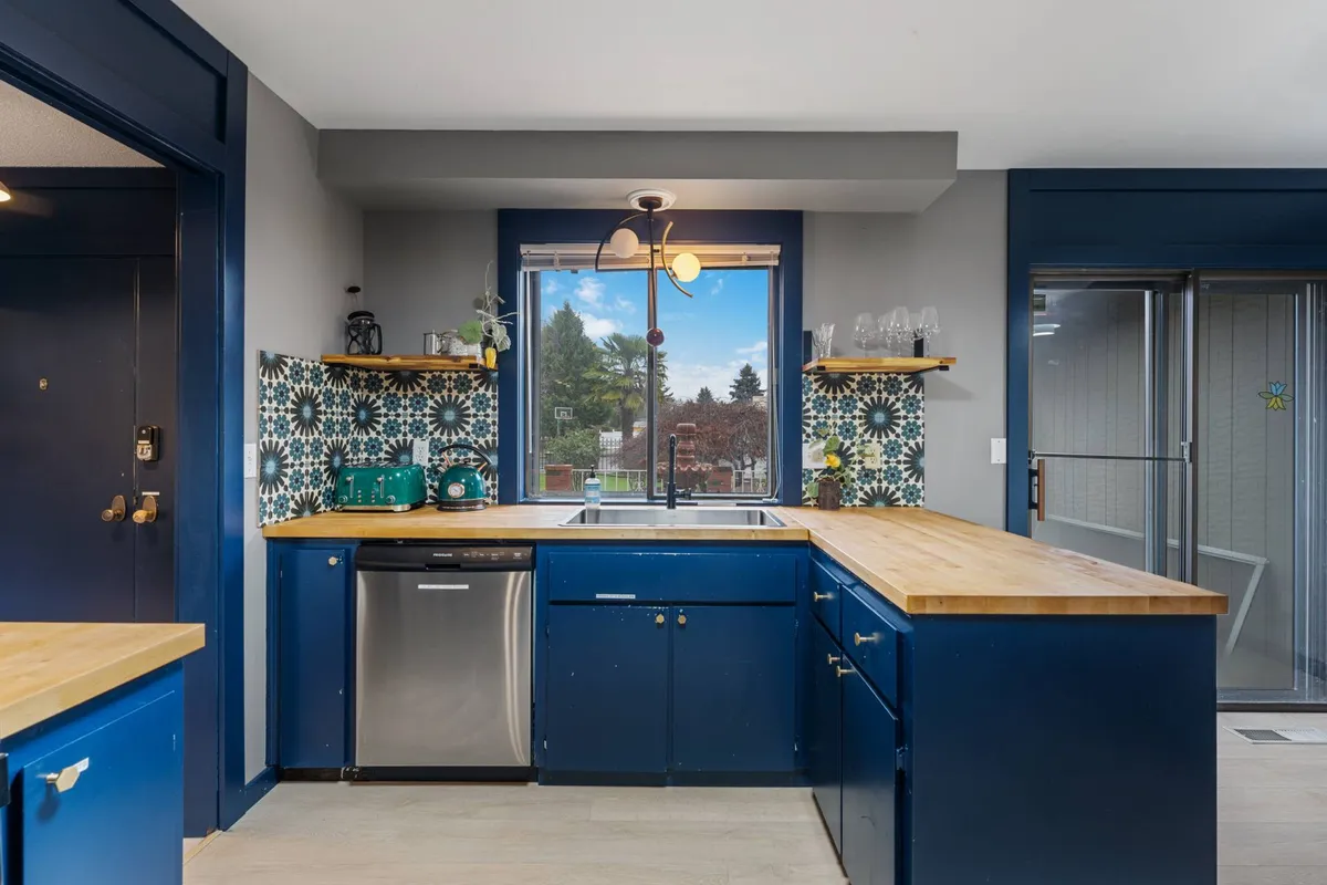 A warm, welcoming kitchen with blue cabinetry, wooden countertops, decorative tiles, and a window view. The space is designed for both cooking and socializing, with visible appliances and homey details.
