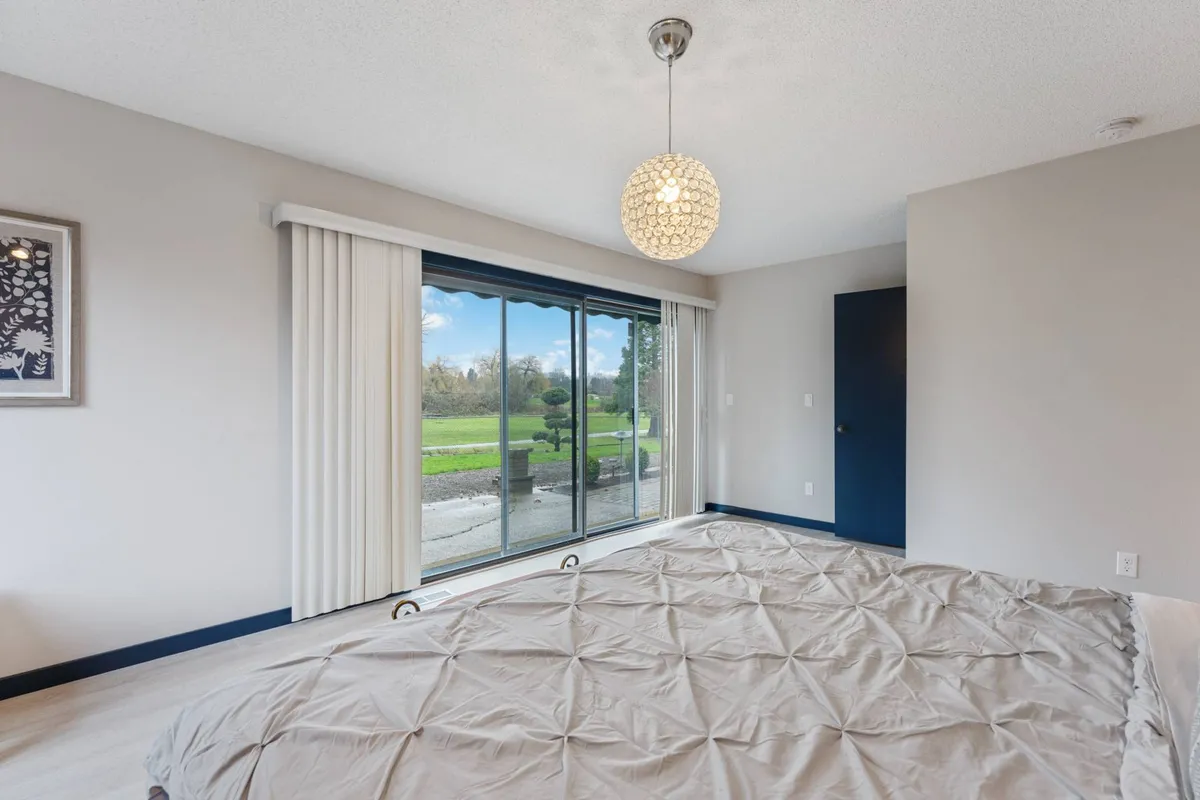 A bright bedroom with a modern light fixture, a bed dressed in a textured bedspread, and a large glass sliding door that lets in abundant natural light and offers a view of the garden. The scene conveys a soothing, welcoming retreat, ideal for extended stays.