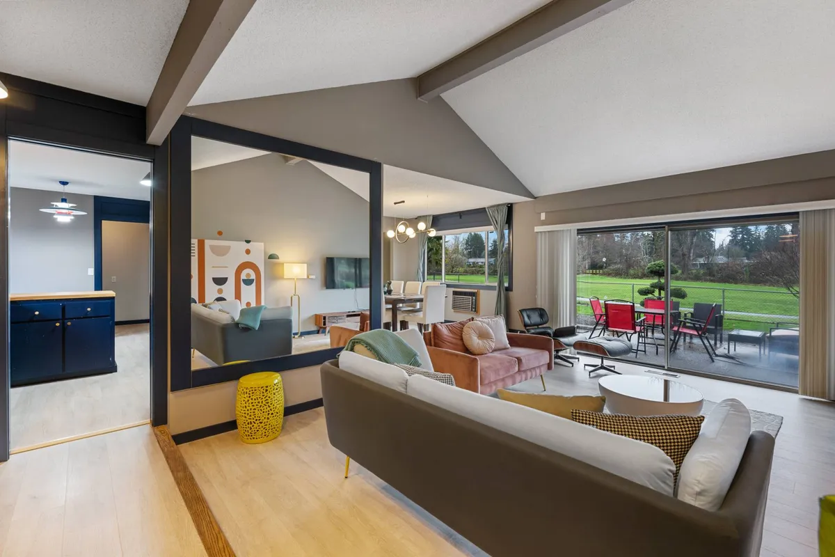 The image showcases a sunlit open-concept living and dining space, featuring a plush sofa facing a large window with views of the outdoors. Colorful decor and a stylish mirror add warmth and character, while a spacious dining area invites group gatherings.